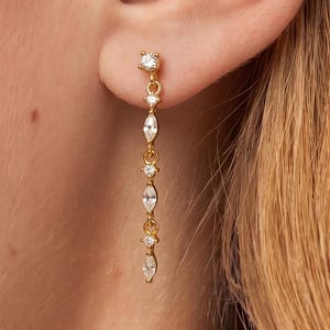 May include: A gold-toned drop earring featuring a vertical chain of alternating round and marquise-cut clear stones. The earring is attached to a small stud post at the top, with the chain dangling downwards, showcasing a series of sparkling white gems.