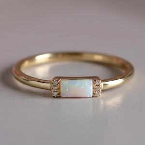 Peut inclure: Une bague en or avec une pierre centrale en opale rectangulaire. De petites pierres claires sont serties de chaque côté de l'opale. La bague est posée sur un fond doux et neutre avec des fleurs roses floues en haut à gauche.