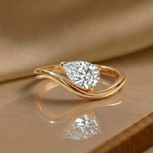 Può includere: Un anello d'oro con un diamante a forma di pera. La fascia dell'anello si curva attorno al diamante, creando un design unico. L'anello è posto su uno sfondo neutro, evidenziando la sua brillantezza ed eleganza. Un bellissimo gioiello.