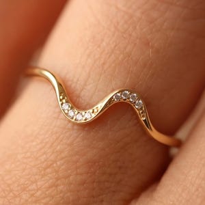 Può includere: Un sottile anello in oro caratterizzato da un delicato design a fascia ondulata o serpentina. La parte superiore dell'onda è accentuata da una serie di piccole pietre chiare e scintillanti incastonate lungo la curva, per un aspetto raffinato.