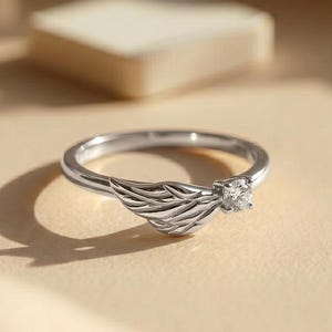 Silver angel wing diamond ring displayed in a luxury brown satin Jewelry box, featuring a detailed wing design beside a round centre stone on a slim polished silver band. A beautiful ring for promise, anniversary, bridal Jewelry, daily wear, angel inspired Jewelry, and fine Jewelry gifting. Perfect for buyers looking for a silver wing ring with clean sparkle and handmade style.