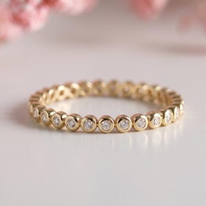 May include: A gold band ring with small, round, clear gemstones set around the entire band. The ring is on a white surface with soft pink flowers in the background.