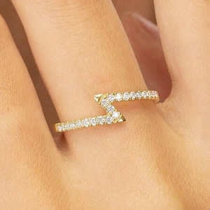 May include: A gold-colored ring with a zig-zag design, encrusted with small, clear gemstones. The ring is worn on a finger, showcasing its delicate and modern aesthetic. The ring's design is a contemporary take on a classic jewelry piece.