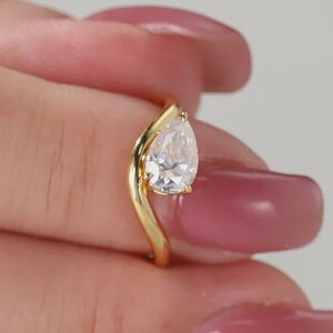 Può includere: Un anello d'oro con una gemma trasparente a forma di pera. La fascia dell'anello è curva, creando un design moderno ed elegante. La gemma è ben incastonata, mettendo in risalto la sua brillantezza. È un gioiello.