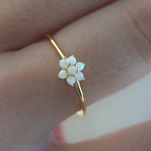 Könnte beinhalten: Ein zarter Goldring mit einem Blumenmuster. Die Blume besteht aus kleinen, weißen Edelsteinen. Der Ring hat ein dünnes, goldenes Band und wird an einem Finger gezeigt.