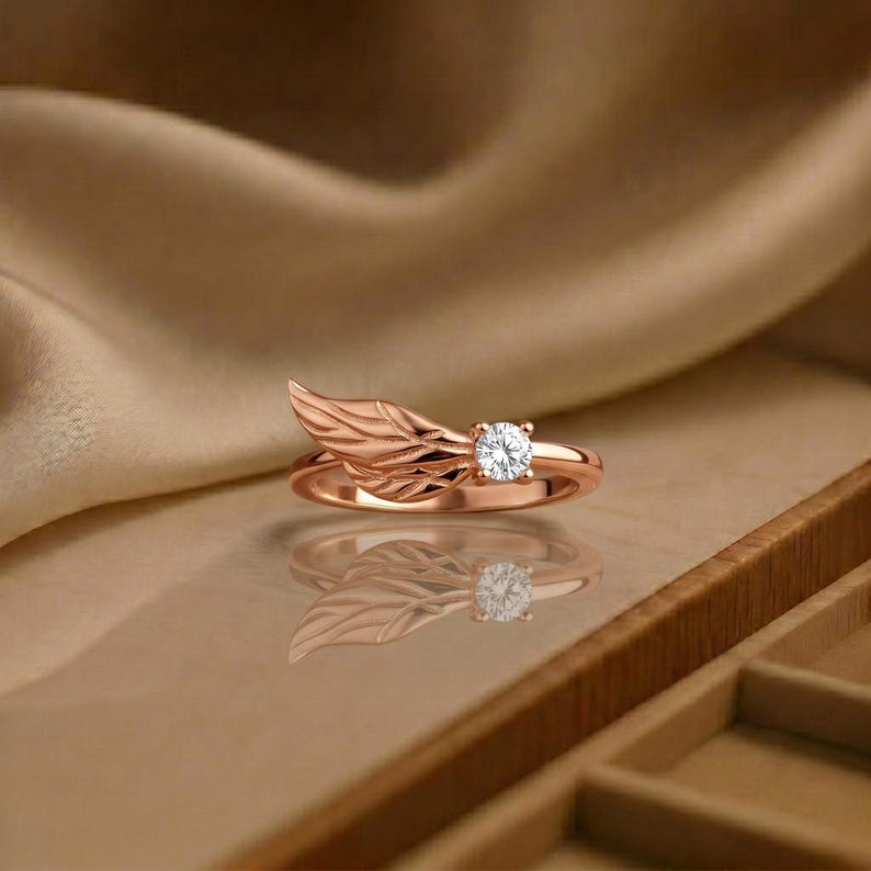Silver angel wing diamond ring displayed in a luxury brown satin Jewelry box, featuring a detailed wing design beside a round centre stone on a slim polished silver band. A beautiful ring for promise, anniversary, bridal Jewelry, daily wear, angel inspired Jewelry, and fine Jewelry gifting. Perfect for buyers looking for a silver wing ring with clean sparkle and handmade style.