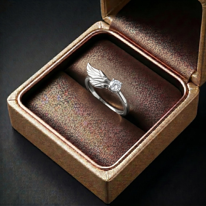 Silver angel wing diamond ring displayed in a luxury brown satin Jewelry box, featuring a detailed wing design beside a round centre stone on a slim polished silver band. A beautiful ring for promise, anniversary, bridal Jewelry, daily wear, angel inspired Jewelry, and fine Jewelry gifting. Perfect for buyers looking for a silver wing ring with clean sparkle and handmade style.