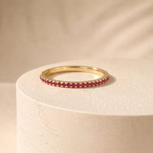 Op de afbeelding: Een dunne gouden ring met een doorlopende rij kleine, ronde, levendige roze-rode edelstenen langs de gehele omtrek. Het metaal heeft een gepolijste afwerking en de stenen zijn uniform van grootte, wat zorgt voor een delicaat, cirkelvormig ontwerp.