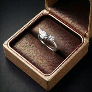 Silver angel wing diamond ring displayed in a luxury brown satin Jewelry box, featuring a detailed wing design beside a round centre stone on a slim polished silver band. A beautiful ring for promise, anniversary, bridal Jewelry, daily wear, angel inspired Jewelry, and fine Jewelry gifting. Perfect for buyers looking for a silver wing ring with clean sparkle and handmade style.