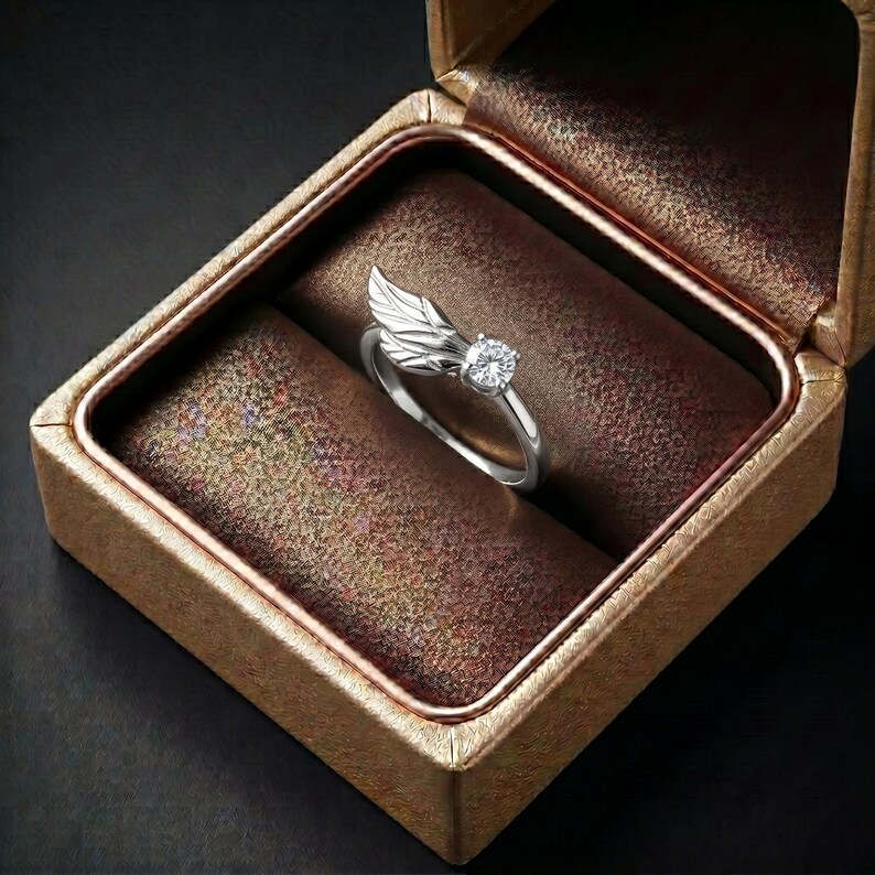 Silver angel wing diamond ring displayed in a luxury brown satin Jewelry box, featuring a detailed wing design beside a round centre stone on a slim polished silver band. A beautiful ring for promise, anniversary, bridal Jewelry, daily wear, angel inspired Jewelry, and fine Jewelry gifting. Perfect for buyers looking for a silver wing ring with clean sparkle and handmade style.