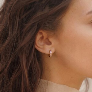 May include: A small gold hoop earring featuring a row of embedded light purple rectangular stones. The earring is secured in the earlobe, showcasing a polished metallic finish and a series of evenly spaced, faceted lavender-colored gemstones.