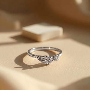 Silver angel wing diamond ring displayed in a luxury brown satin Jewelry box, featuring a detailed wing design beside a round centre stone on a slim polished silver band. A beautiful ring for promise, anniversary, bridal Jewelry, daily wear, angel inspired Jewelry, and fine Jewelry gifting. Perfect for buyers looking for a silver wing ring with clean sparkle and handmade style.