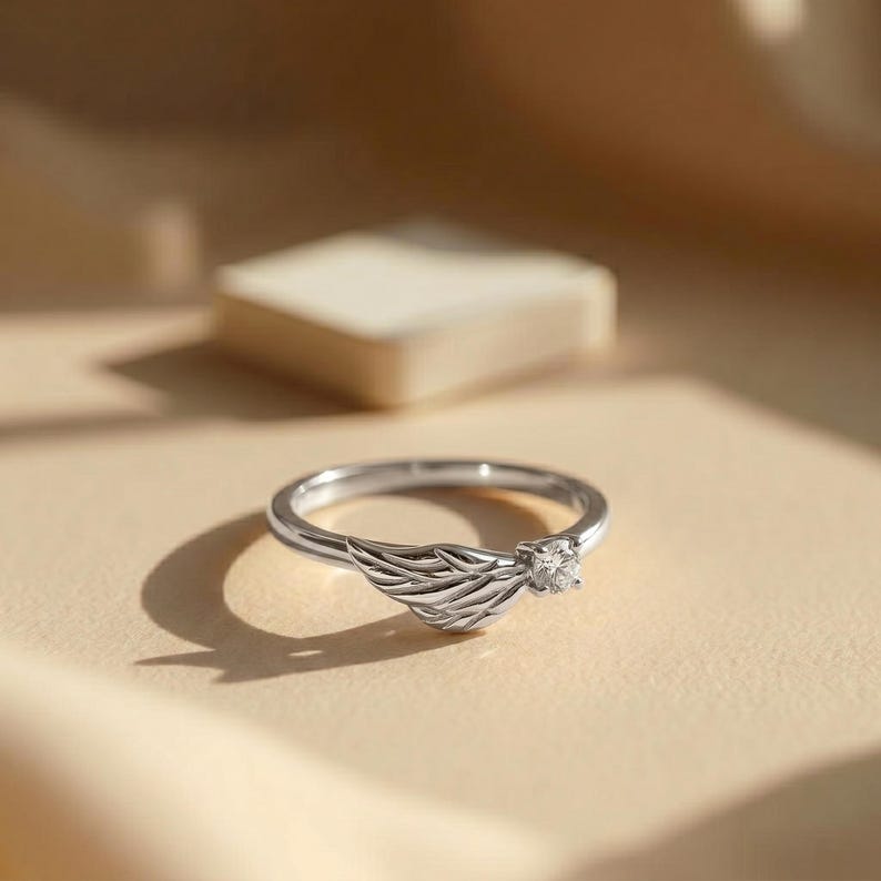Silver angel wing diamond ring displayed in a luxury brown satin Jewelry box, featuring a detailed wing design beside a round centre stone on a slim polished silver band. A beautiful ring for promise, anniversary, bridal Jewelry, daily wear, angel inspired Jewelry, and fine Jewelry gifting. Perfect for buyers looking for a silver wing ring with clean sparkle and handmade style.