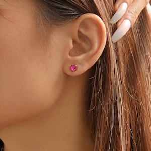 Könnte beinhalten: Ein kleiner Ohrstecker mit einem leuchtend pinken, herzförmigen Edelstein in einer goldfarbenen Krappenfassung, der im Ohrläppchen sitzt. Das Design ist schlicht und kompakt und betont die facettierte Oberfläche des pinken Steins.