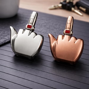Op de afbeelding: Twee novelty aanstekers in de vorm van handen met de middelvinger uitgestoken. Eén is zilverkleurig, de andere koperkleurig. Elke aansteker heeft een rode rechthoekige accent op de vinger. De aanstekers liggen op een zwart, geribbeld oppervlak.