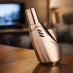 May include: A gold-coloured JOBON lighter with a sleek, modern design. The lighter features a cylindrical top, a curved body with three small vents, and the brand name "JOBON" printed on the side. It is resting on a wooden surface.