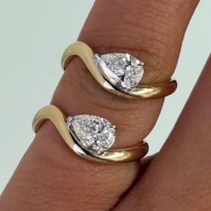 May include: Two gold rings, each featuring a pear-cut diamond in a swirling setting. The rings are shown on a finger. The diamonds are clear and reflect light, set against a pale background.
