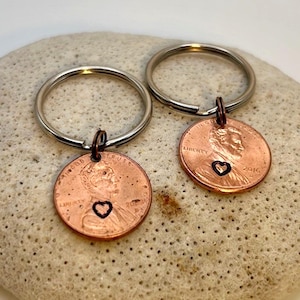 May include: Two copper penny keychains. Each penny has a heart cutout and is attached to a silver keyring. The pennies are dated 2016 and feature the profile of Abraham Lincoln. The keychains are displayed on a textured, off-white surface.