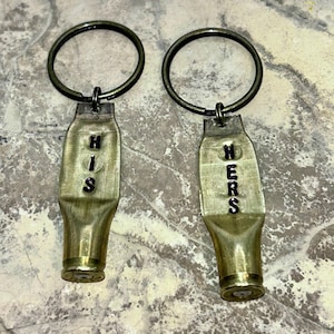 May include: Two brass keychains, each crafted from a bullet casing. One keychain reads "HIS" and the other reads "HERS" in black lettering. Both keychains have a bronze-colored ring.