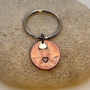 May include: A copper penny keychain with a small silver heart charm and a heart-shaped cutout. The penny is attached to a silver keyring. The penny shows the year 2008.