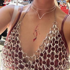 May include: A silver-toned chainmail top featuring a repeating pattern of interlocking metal rings, worn over a dark mauve garment. A double-layered silver V-shaped choker necklace rests at the neck, and a small red and black temporary tattoo is visible.