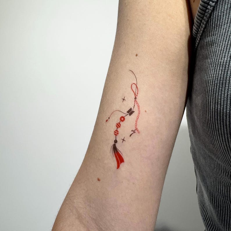 May include: A temporary tattoo design featuring a delicate red string element with three circular red beads, a small black arrow icon, the number seven, and a red tassel at the bottom, accented by thin black lines and small cross symbols.