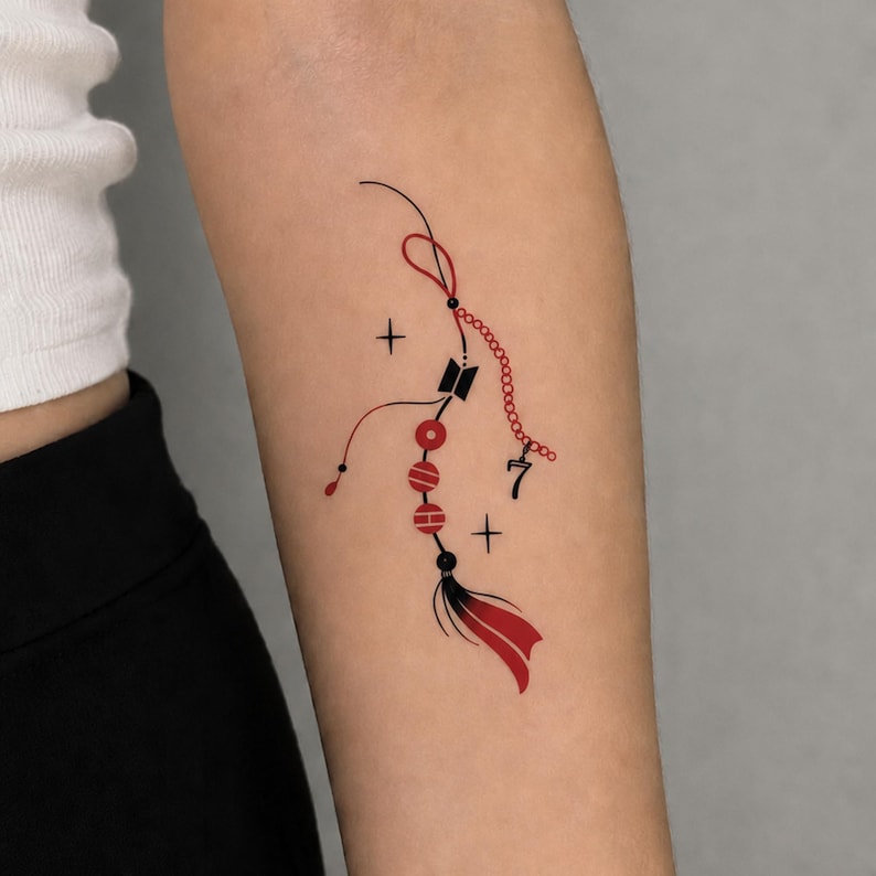 May include: A temporary tattoo design featuring a red and black decorative charm with a central black logo, a dangling number seven, three red circular beads, and a red and black tassel, accented by two small black four-pointed star symbols.