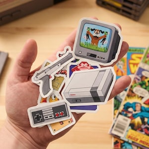 May include: A collection of retro video game-themed stickers featuring a gray console, controller, and pixelated graphics. The stickers include a TV screen with a hunting dog, a gun, and a red-haired character. A Nintendo console and game cartridges are in the background.