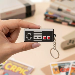 May include: Black and white keychain shaped like a classic video game controller, with red buttons. The keychain has a silver chain and ring. Other retro gaming items are in the background.