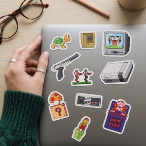 May include: A collection of colorful, pixelated video game-themed stickers on a laptop. The stickers include a turtle, a gun, a television, a game console, a mushroom with a question mark, a controller, and a "Game Over" sign.