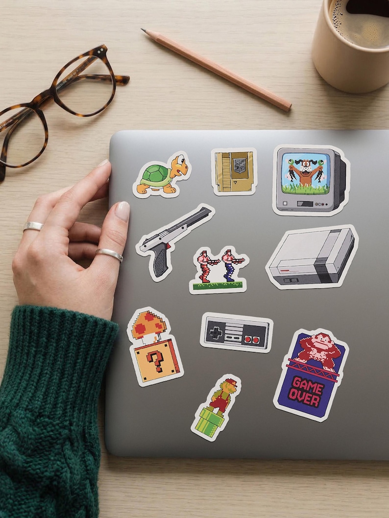 May include: A collection of colorful, pixelated video game-themed stickers on a laptop. The stickers include a turtle, a gun, a television, a game console, a mushroom with a question mark, a controller, and a "Game Over" sign.