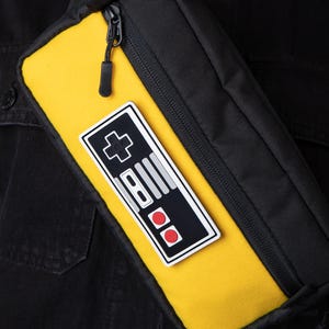 Könnte beinhalten: Schwarze und gelbe Bauchtasche mit einem Retro-Videospiel-Controller-Patch. Die Tasche hat einen schwarzen Reißverschluss und einen schwarzen Riemen. Der Controller-Patch ist schwarz, weiß und rot.