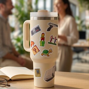 May include: A cream-colored insulated tumbler with a handle and lid, adorned with colorful stickers of retro video game characters and icons. The stickers include a controller, a gun, and a question mark.