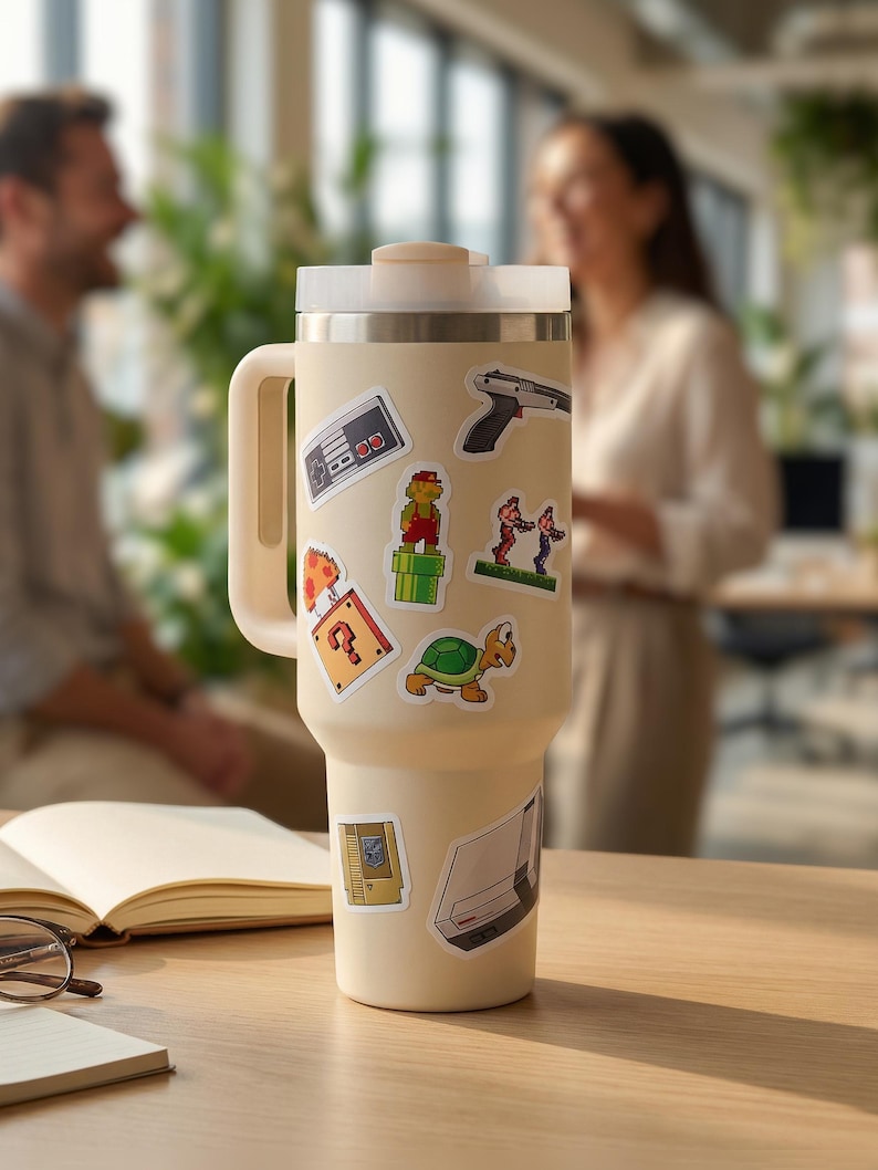 May include: A cream-colored insulated tumbler with a handle and lid, adorned with colorful stickers of retro video game characters and icons. The stickers include a controller, a gun, and a question mark.