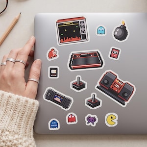May include: A collection of pixelated retro video game stickers, including a television, game console, joystick, and various game characters like Pac-Man and ghosts. The stickers are placed on a silver laptop.