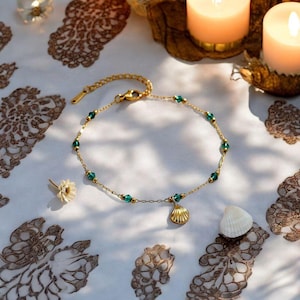 Puede incluir: Una tobillera de cadena dorada con pequeñas cuentas facetadas de color verde esmeralda espaciadas uniformemente. Un dije de concha marina de color dorado cuelga del centro de la cadena, que incluye un cierre de mosquetón y una extensión.