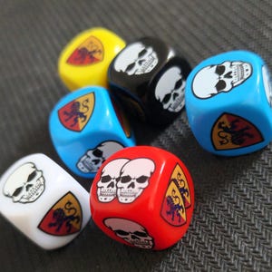 Pode incluir: Um conjunto de dados de seis lados em várias cores, incluindo vermelho, azul, branco, preto e amarelo. Cada dado apresenta gráficos impressos de crânios humanos brancos e emblemas em forma de escudo com um leão preto sobre fundo amarelo e vermelho.