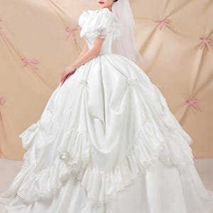 K&ouml;nnte beinhalten: Ein wei&szlig;es Ballkleid mit Puff&auml;rmeln, einem eng anliegenden Oberteil und einem volumin&ouml;sen Rock mit geschichteten R&uuml;schen und gerafften Stoffakzenten. Das Kleid wird mit einem transparenten wei&szlig;en Schleier kombiniert, der &uuml;ber Kopf und Schultern f&auml;llt.