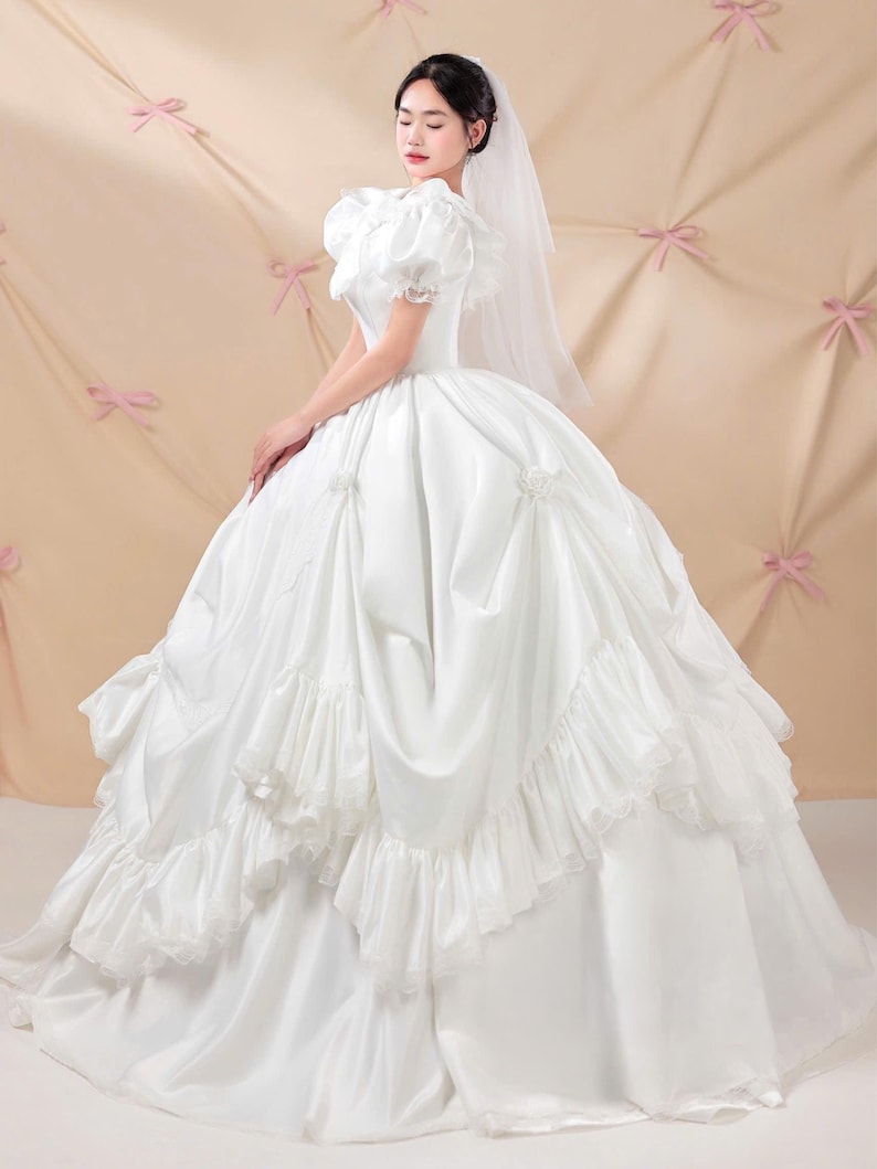 K&ouml;nnte beinhalten: Ein wei&szlig;es Ballkleid mit Puff&auml;rmeln, einem eng anliegenden Oberteil und einem volumin&ouml;sen Rock mit geschichteten R&uuml;schen und gerafften Stoffakzenten. Das Kleid wird mit einem transparenten wei&szlig;en Schleier kombiniert, der &uuml;ber Kopf und Schultern f&auml;llt.