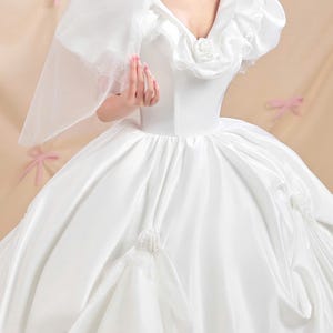 K&ouml;nnte beinhalten: Ein wei&szlig;es Ballkleid mit V-Ausschnitt, R&uuml;schenbesatz und einer zentralen Stoffrose. Das Kleid hat Puff&auml;rmel, einen volumin&ouml;sen Rock mit gerafften Stoffakzenten und einen transparenten Schleier, der am Oberteil befestigt ist und eine Silhouette bildet.