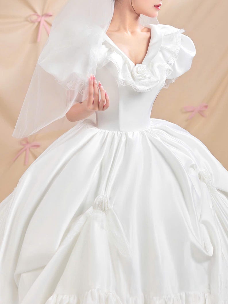 K&ouml;nnte beinhalten: Ein wei&szlig;es Ballkleid mit V-Ausschnitt, R&uuml;schenbesatz und einer zentralen Stoffrose. Das Kleid hat Puff&auml;rmel, einen volumin&ouml;sen Rock mit gerafften Stoffakzenten und einen transparenten Schleier, der am Oberteil befestigt ist und eine Silhouette bildet.