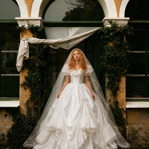 K&ouml;nnte beinhalten: Ein wei&szlig;es Ballkleid aus Satin mit Puff&auml;rmeln, einem figurbetonten Oberteil und einem volumin&ouml;sen Rock mit gerafften Stofflagen und Spitzenbesatz. Ein langer, transparenter Schleier mit Spitzenrand liegt auf dem Kopf und f&auml;llt bis zum Boden.