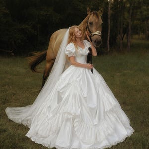 K&ouml;nnte beinhalten: Ein wei&szlig;es, bodenlanges Ballkleid mit Puff&auml;rmeln, herzf&ouml;rmigem Ausschnitt und einem volumin&ouml;sen Rock mit gestuften R&uuml;schen und Spitzenbesatz. Ein langer, transparenter wei&szlig;er Schleier ist an der R&uuml;ckseite des Kleides befestigt.