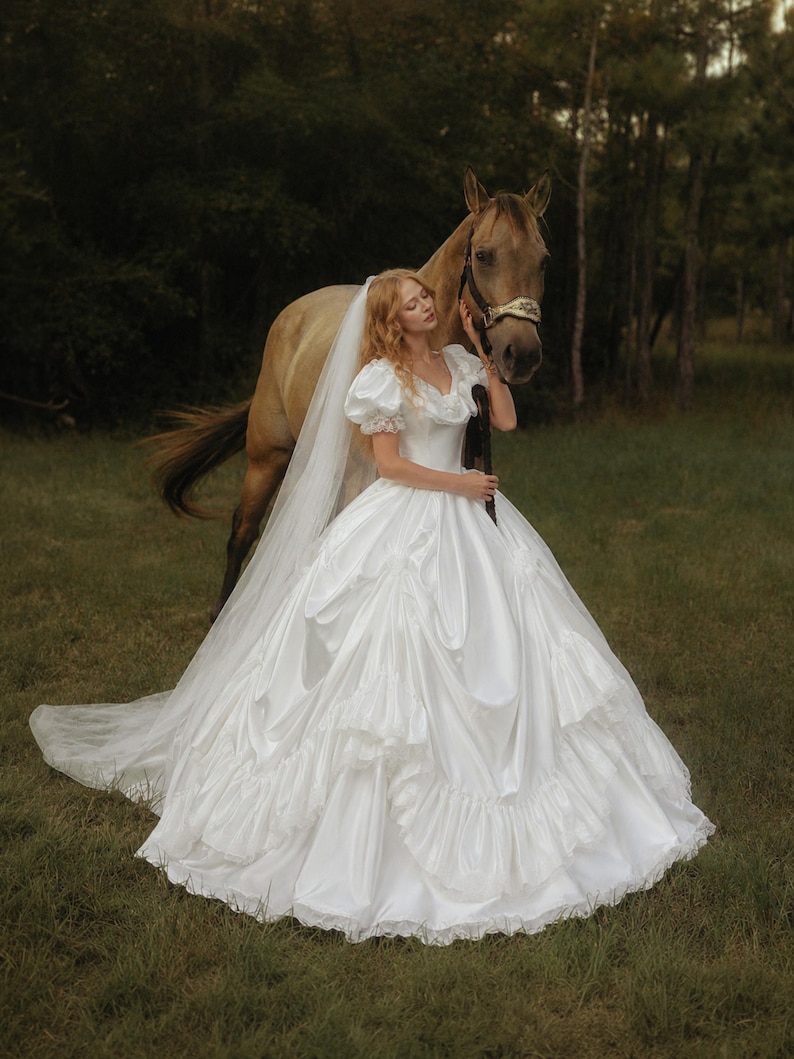 K&ouml;nnte beinhalten: Ein wei&szlig;es, bodenlanges Ballkleid mit Puff&auml;rmeln, herzf&ouml;rmigem Ausschnitt und einem volumin&ouml;sen Rock mit gestuften R&uuml;schen und Spitzenbesatz. Ein langer, transparenter wei&szlig;er Schleier ist an der R&uuml;ckseite des Kleides befestigt.