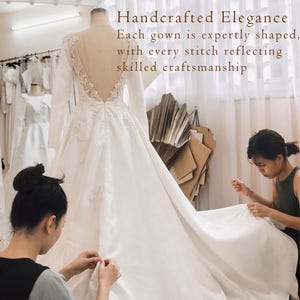 K&ouml;nnte beinhalten: Ein wei&szlig;es lang&auml;rmeliges Kleid mit tiefem V-Ausschnitt am R&uuml;cken, transparenten Eins&auml;tzen und floralen Spitzenapplikationen. Der Text lautet: Handcrafted Elegance. Each gown is expertly shaped, with every stitch reflecting skilled craftsmanship.
