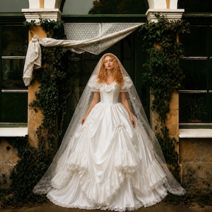 K&ouml;nnte beinhalten: Ein wei&szlig;es Ballkleid mit Puff&auml;rmeln, herzf&ouml;rmigem Ausschnitt und einem volumin&ouml;sen Rock mit geschichteten R&uuml;schen und gerafften Stoffdetails. Das Kleid wird mit einem langen, transparenten, spitzenbesetzten Schleier &uuml;ber den Schultern kombiniert.