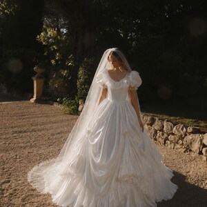 K&ouml;nnte beinhalten: Ein bodenlanges wei&szlig;es Brautkleid mit weitem Ausschnitt, Puff&auml;rmeln und einem volumin&ouml;sen Rock mit gerafften Stoffdetails und R&uuml;schenbesatz. Das Kleid wird mit einem langen, transparenten, bodenlangen Schleier aus feinem T&uuml;llstoff kombiniert.