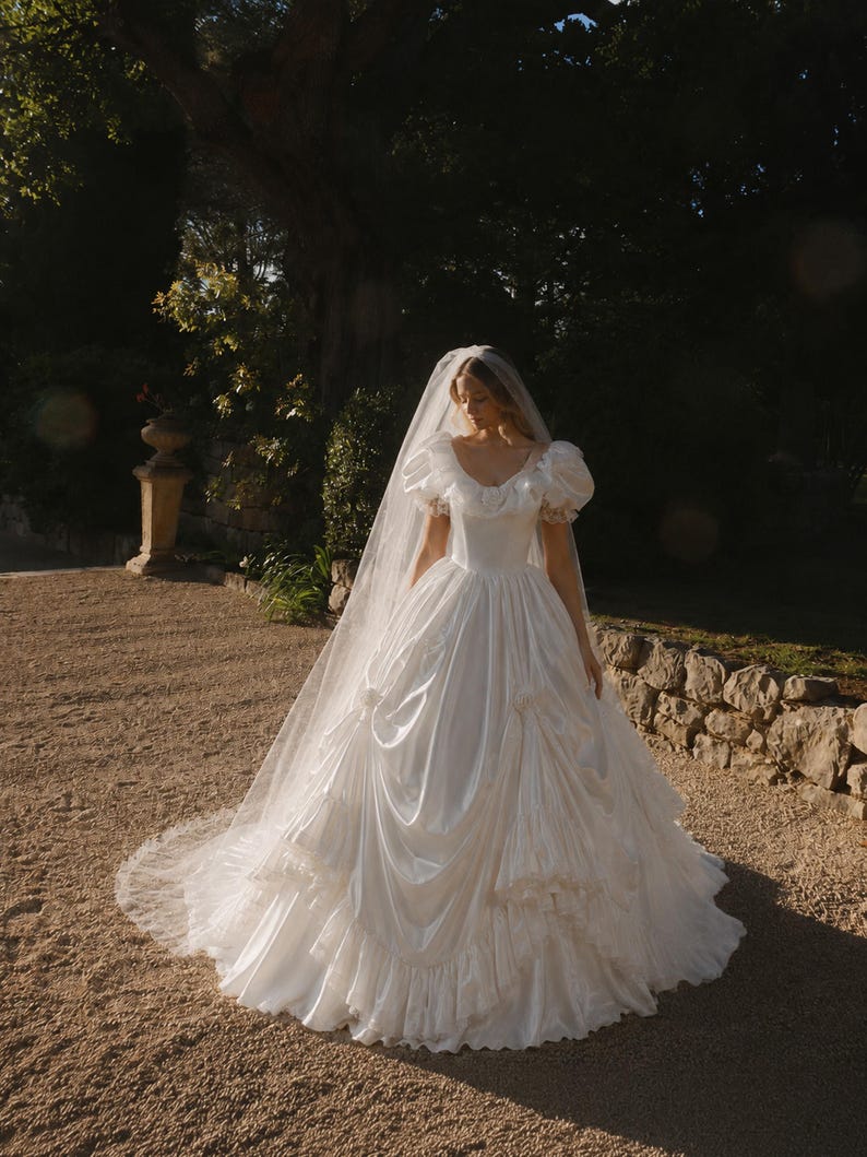 K&ouml;nnte beinhalten: Ein bodenlanges wei&szlig;es Brautkleid mit weitem Ausschnitt, Puff&auml;rmeln und einem volumin&ouml;sen Rock mit gerafften Stoffdetails und R&uuml;schenbesatz. Das Kleid wird mit einem langen, transparenten, bodenlangen Schleier aus feinem T&uuml;llstoff kombiniert.
