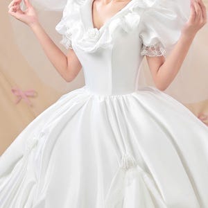 K&ouml;nnte beinhalten: Ein wei&szlig;es Ballkleid aus Satin mit V-Ausschnitt und R&uuml;schenbesatz, kurzen Puff&auml;rmeln mit Spitzenkanten und einem weiten Rock, der mit Stoffrosen verziert ist. Das Kleid wird mit einem transparenten wei&szlig;en Schleier &uuml;ber den Schultern kombiniert.