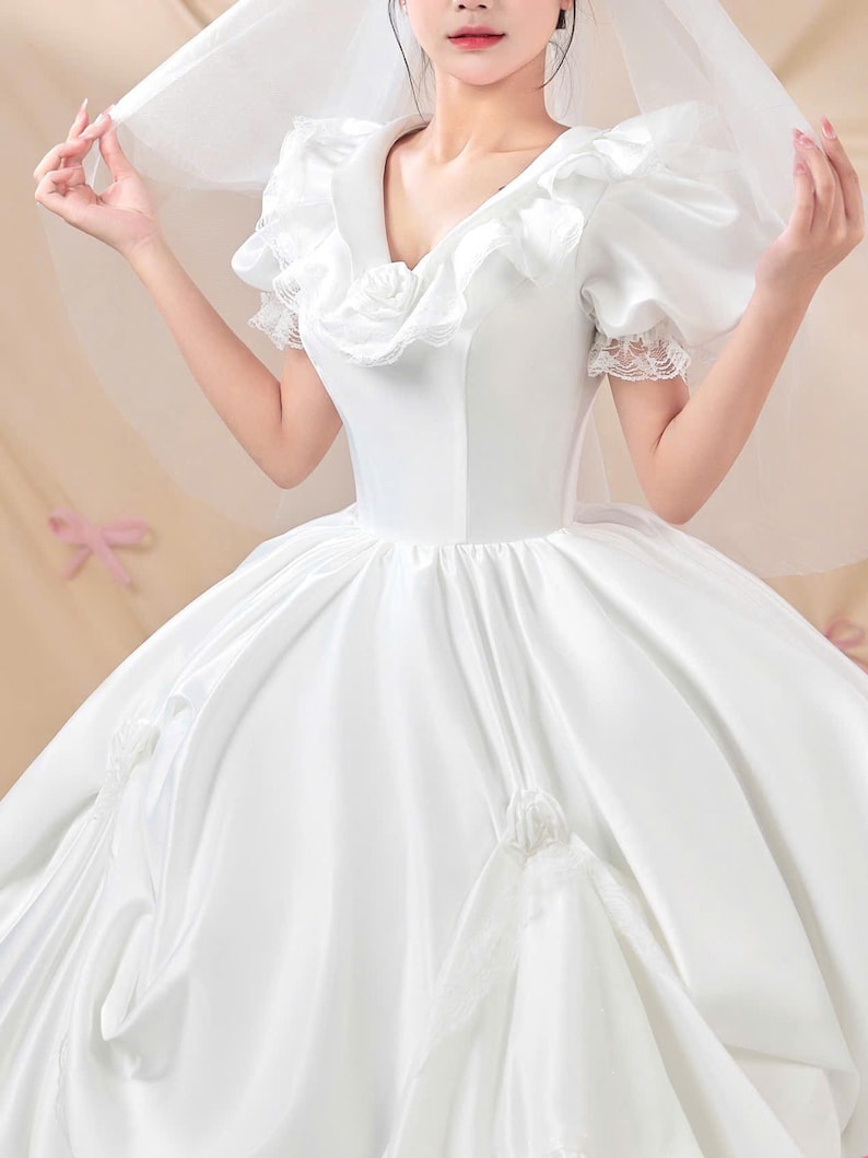 K&ouml;nnte beinhalten: Ein wei&szlig;es Ballkleid aus Satin mit V-Ausschnitt und R&uuml;schenbesatz, kurzen Puff&auml;rmeln mit Spitzenkanten und einem weiten Rock, der mit Stoffrosen verziert ist. Das Kleid wird mit einem transparenten wei&szlig;en Schleier &uuml;ber den Schultern kombiniert.